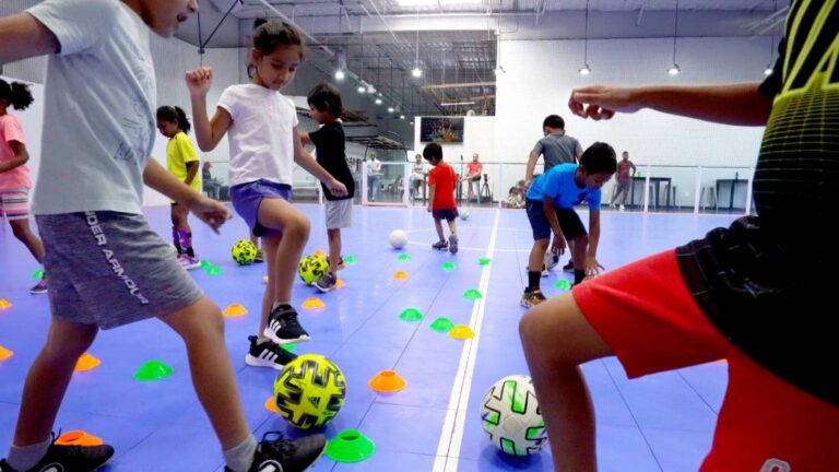 Many young children using their feet to dribble a soccer ball between cones