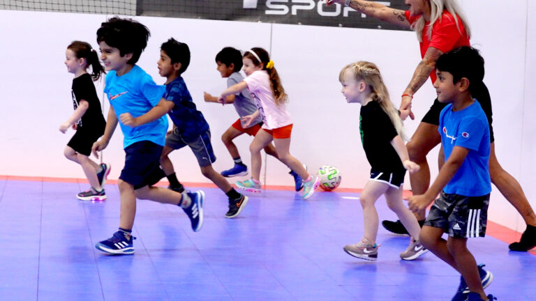 Kids running playing soccer with an instructor on an indoor soccer field.