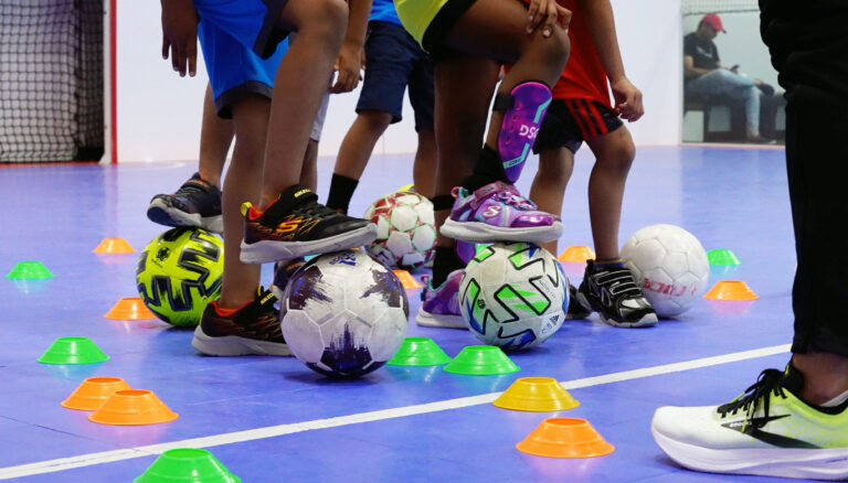 Waist down view of several children with one foot on a soccer ball. Several soccer balls and colorful cones are seen in the image.