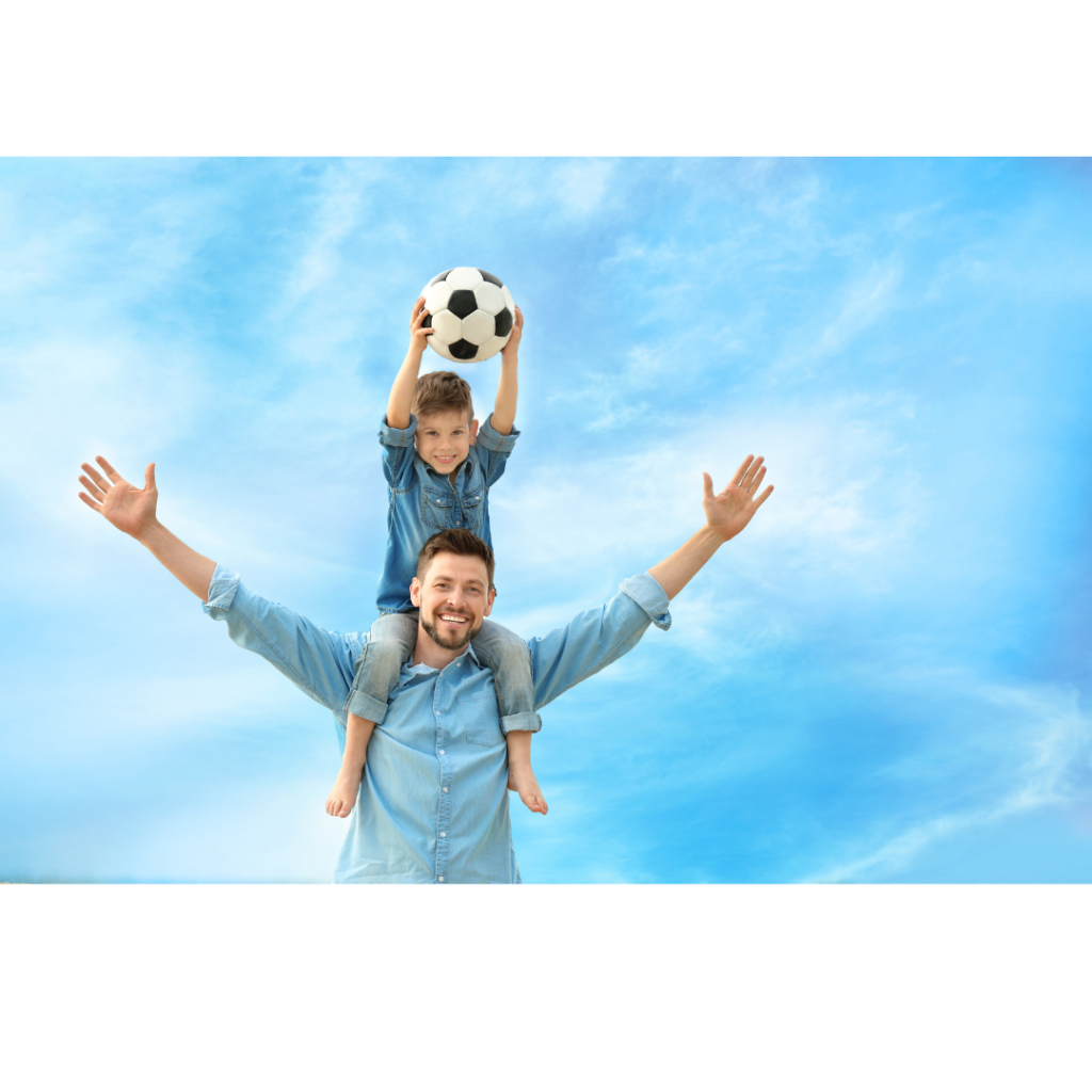 Happy dad holds son on his shoulders, who is holding a soccer ball above his head.
