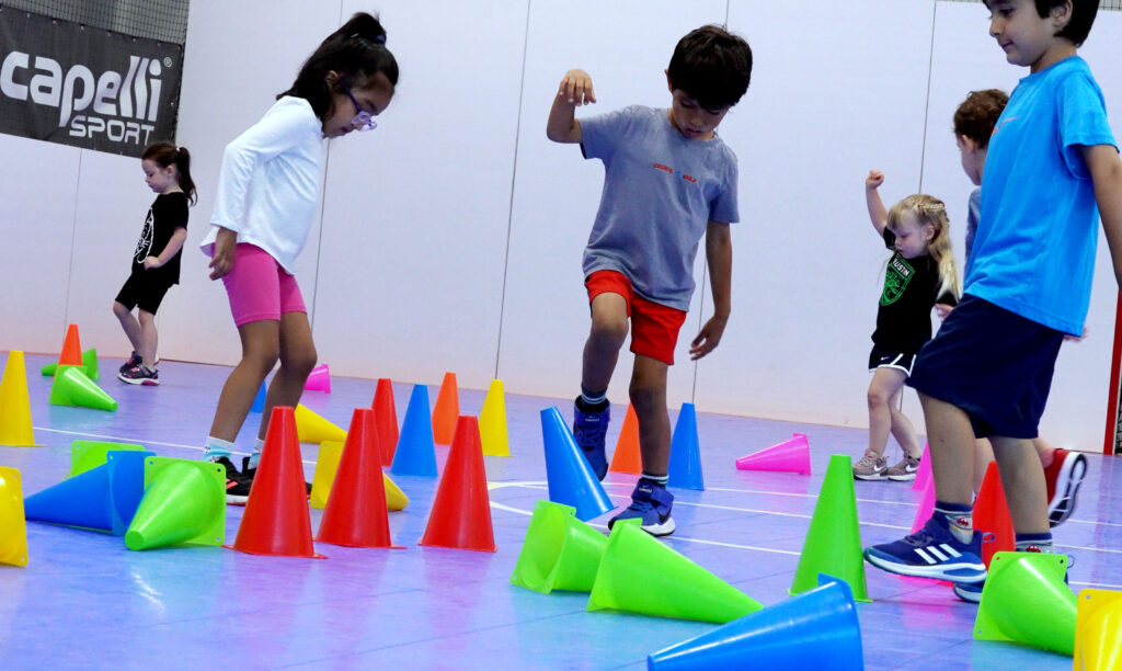 Young kids are seen doing a soccer drill with multi colored cones.